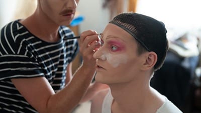 Eine Person wird von einer Visagistin mit auffälligem Make-up geschminkt – Symbolbild für Drag