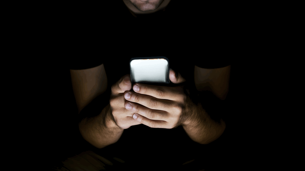 Hände eines Menschen, der in dunkler Umgebung auf ein Smartphone schaut