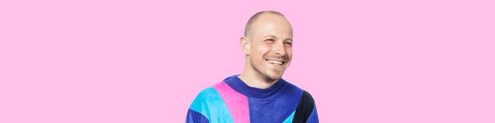 Robi on a pink background - intersex activist in a blue and black shirt with rainbow elements