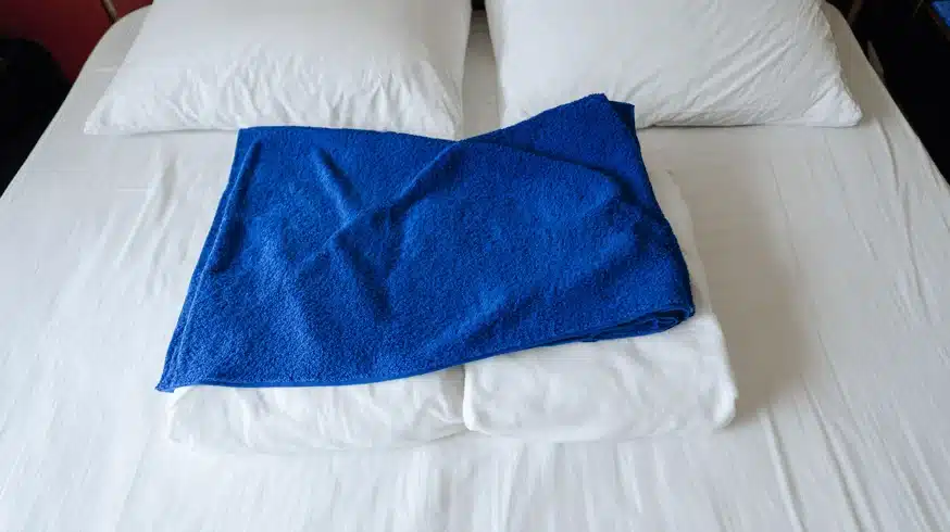 Gefaltetes blaues Handtuch liegt auf einem frisch gemachten Bett mit weißen Laken.