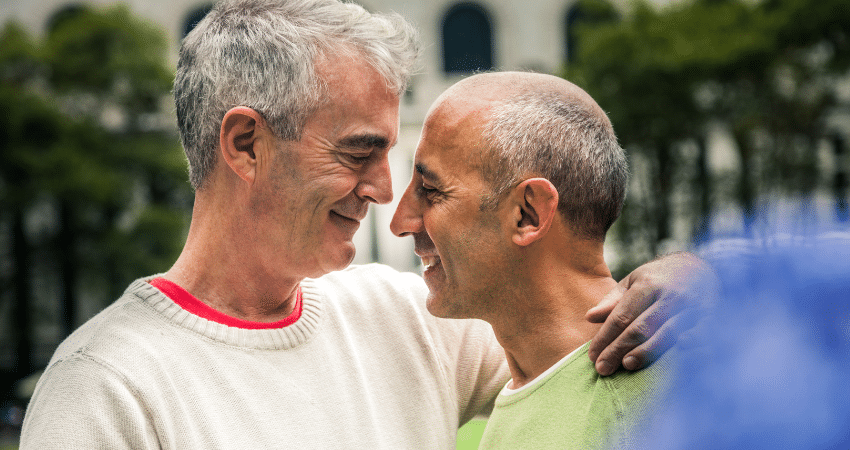 Two older men look at each other lovingly and embrace tenderly.